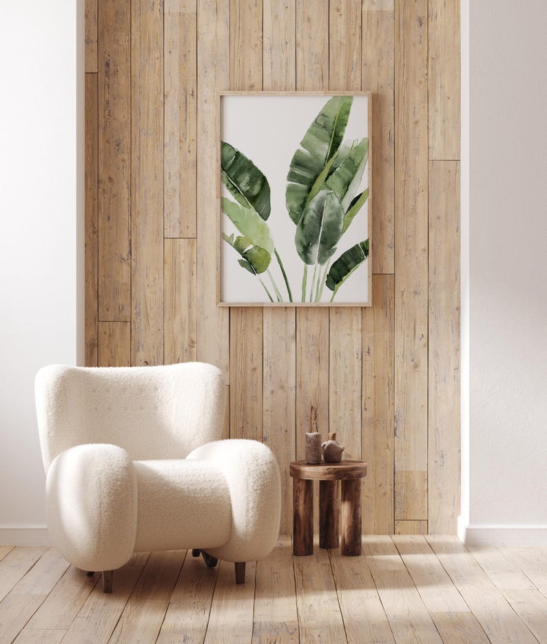 May include: A white fluffy armchair with a wooden side table and a framed print of green tropical leaves on a wood panel wall.