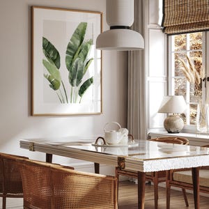May include: A dining room with a white table and chairs with woven cane seats. The table has a white marble top and a glass insert. A white pendant light hangs over the table. A framed print of green banana leaves hangs on the wall.