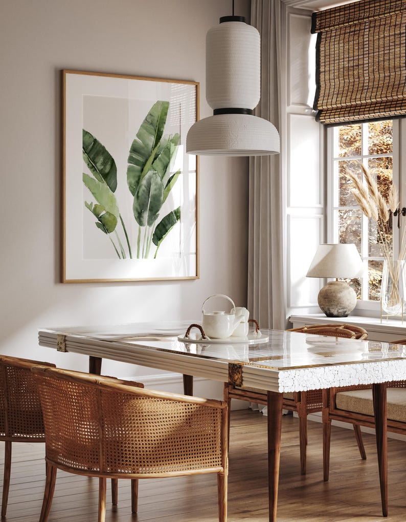 May include: A dining room with a white table and chairs with woven cane seats. The table has a white marble top and a glass insert. A white pendant light hangs over the table. A framed print of green banana leaves hangs on the wall.