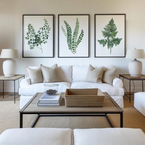 May include: A white sofa with three throw pillows sits in front of a white wall with three framed botanical prints of green ferns. A coffee table with a woven basket and books sits in front of the sofa. Two end tables with lamps are on either side of the sofa.