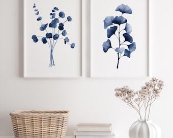 Può includere: Due stampe ad acquerello incorniciate di illustrazioni botaniche blu. La stampa a sinistra presenta un bouquet di piccoli fiori blu. La stampa a destra presenta un ramo di foglie blu. Le stampe sono appese a una parete bianca sopra uno scaffale bianco con un cesto di vimini, libri e un vaso bianco con fiori secchi.