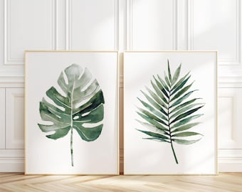 Pode incluir: Duas impressões de aquarela emolduradas de folhas tropicais verdes. A impressão à esquerda apresenta uma grande folha de monstera, enquanto a impressão à direita apresenta uma pequena folha de palmeira.