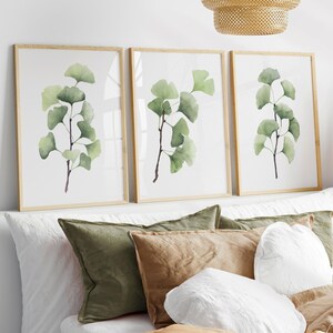 Könnte beinhalten: Drei gerahmte Kunstdrucke mit Aquarellillustrationen von Ginkgoblättern in verschiedenen Grüntönen. Die Drucke haben hellbraune Holzrahmen und werden über einem Bett mit Kissen in Grün-, Braun- und Weißtönen ausgestellt.