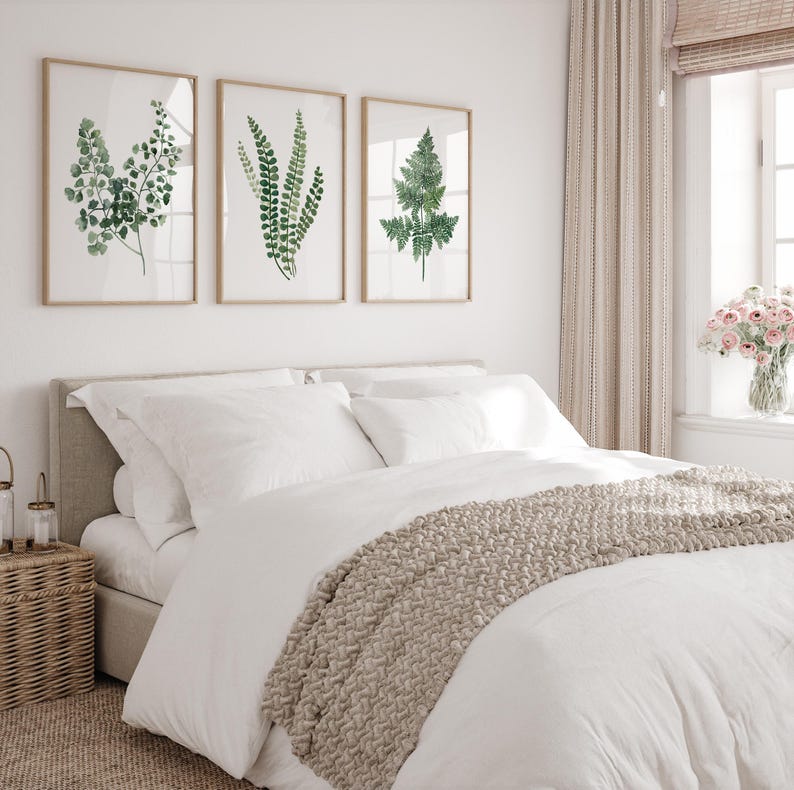 May include: A white bed with a beige knitted throw blanket and three framed botanical prints of ferns on a white wall. The prints are in a simple, modern style.
