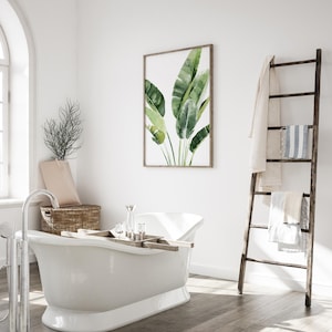 May include: A white freestanding bathtub with a wooden tray and clear glass jars on top. A framed watercolor painting of green banana leaves hangs on the wall behind the bathtub. A wooden ladder with towels hanging on it is in the corner of the room.