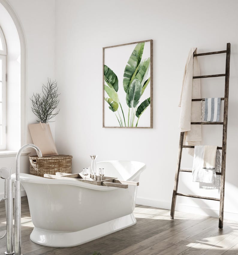 May include: A white freestanding bathtub with a wooden tray and clear glass jars on top. A framed watercolor painting of green banana leaves hangs on the wall behind the bathtub. A wooden ladder with towels hanging on it is in the corner of the room.