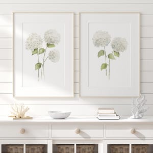 以下が含まれることがあります： 緑の葉を持つ白いアジサイの花の水彩画イラストが描かれた、2枚の縦型ウォールアートプリント。各プリントは明るい色の木製フレームに収められており、白い背景に植物のボタニカルアートが表現されています。