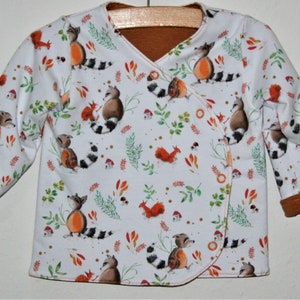 Könnte beinhalten: Weißer Baby-Pullover mit einem braunen und weißen Waschbär-Muster. Der Pullover hat einen braunen gerippten Kragen und Bündchen.