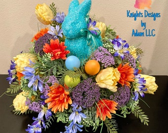 May include: A turquoise glitter bunny sits in a spring floral arrangement. The arrangement includes yellow tulips, orange and red chrysanthemums, purple flowers, and blue flowers. The arrangement is surrounded by green foliage and pastel colored Easter eggs.