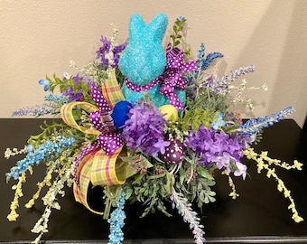 May include: A blue glitter bunny sits in the center of a spring floral arrangement. The arrangement features purple flowers, green foliage, and yellow and purple speckled eggs. A purple and white polka dot ribbon is tied around the bunny's neck.