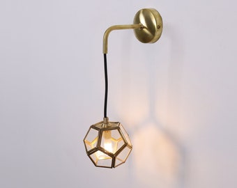 May include: A gold wall sconce with a geometric glass shade. The sconce has a black cord and is mounted on a white wall.