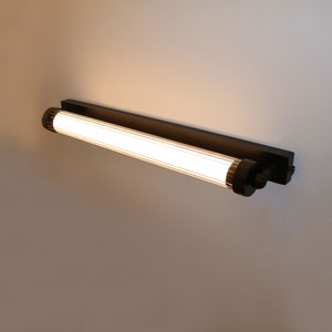 以下が含まれることがあります： 白いすりガラスのチューブが付いた黒い壁掛け照明器具。照明が点灯しており、部屋を照らしています。