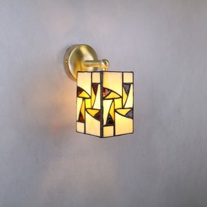 May include: A brass wall sconce with a square stained glass shade. The shade features a geometric pattern of yellow, brown, and white glass pieces.