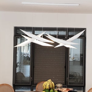 May include: A white modern chandelier with multiple geometric shapes hangs from the ceiling in a dining room. The chandelier is suspended over a table with a bowl of fruit on it.