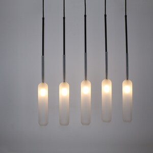 May include: Five frosted glass pendant lights hang from black cords against a white wall. The lights are cylindrical in shape and emit a warm, white glow.