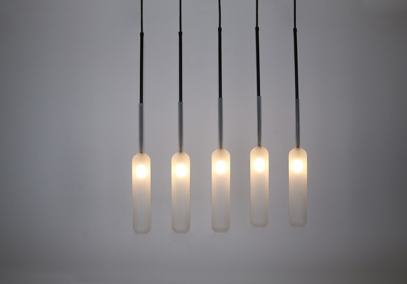 May include: Five frosted glass pendant lights hang from black cords against a white wall. The lights are cylindrical in shape and emit a warm, white glow.