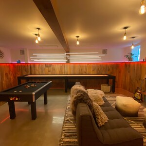 May include: A game room with a pool table, a shuffleboard table, a brown couch, and a rug. The walls are wood paneling with red LED lights.