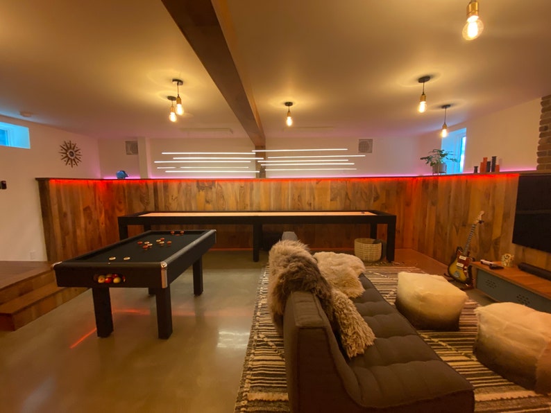May include: A game room with a pool table, a shuffleboard table, a brown couch, and a rug. The walls are wood paneling with red LED lights.