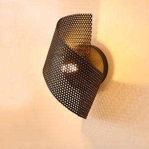 May include: A black metal wall sconce with a perforated shade. The shade is curved and has a light bulb inside.