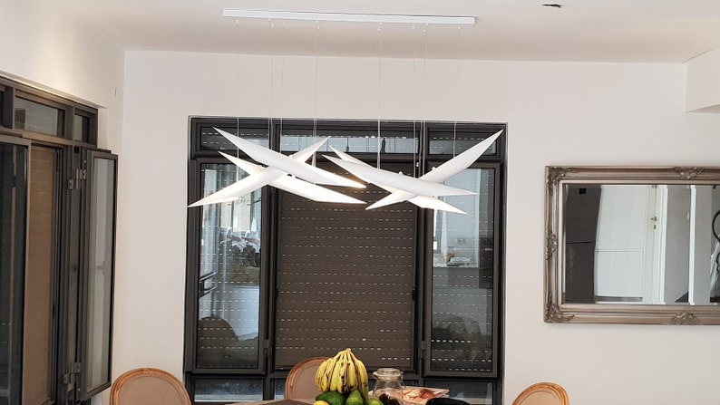 May include: A white modern chandelier with multiple geometric shapes hangs from the ceiling in a dining room. The chandelier is suspended over a table with a bowl of fruit on it.