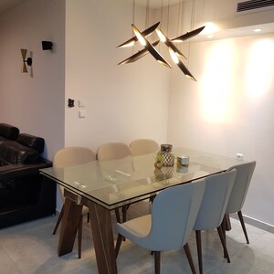 May include: A glass dining table with a wooden base and six beige chairs. The table has a modern design with a clear glass top and a sleek wooden frame. A modern light fixture hangs above the table, adding a touch of contemporary style to the dining room.