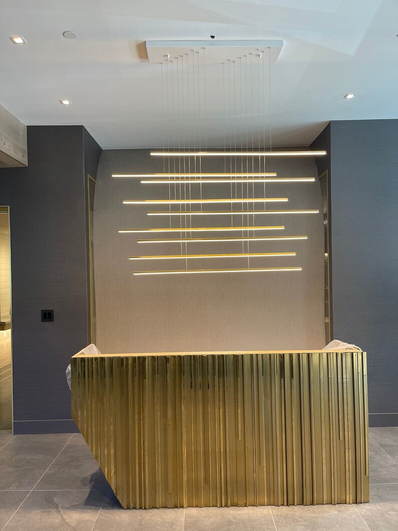 May include: A gold-colored reception desk with vertical grooves, illuminated by a modern light fixture with multiple horizontal bars.