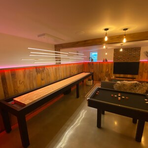 May include: A game room with a wooden shuffleboard table and a black pool table. The walls are wood paneling with red LED lights. There are several hanging light fixtures with Edison bulbs.