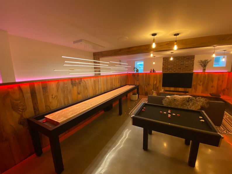 May include: A game room with a wooden shuffleboard table and a black pool table. The walls are wood paneling with red LED lights. There are several hanging light fixtures with Edison bulbs.