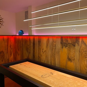 May include: A wooden shuffleboard table with a black frame, set up in a game room with a wood-paneled wall and red and pink LED lights.