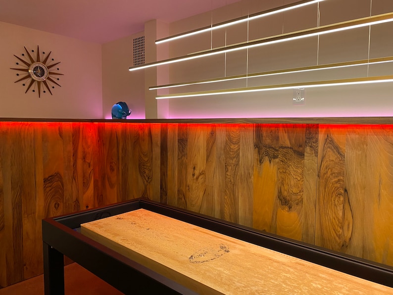 May include: A wooden shuffleboard table with a black frame, set up in a game room with a wood-paneled wall and red and pink LED lights.