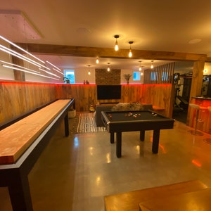 May include: A game room with a wooden shuffleboard table, a pool table, and a bar area with stools. The walls are wood paneling with red LED lights. The room has a modern, industrial feel.