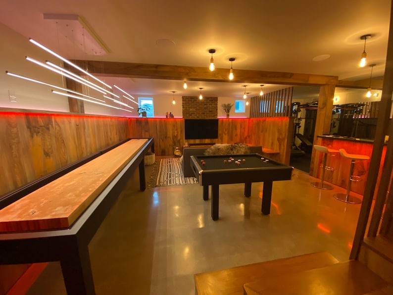 May include: A game room with a wooden shuffleboard table, a pool table, and a bar area with stools. The walls are wood paneling with red LED lights. The room has a modern, industrial feel.