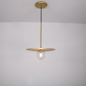 May include: A gold pendant light with a round glass shade and a flat, circular canopy. The light is hanging from a black cord.