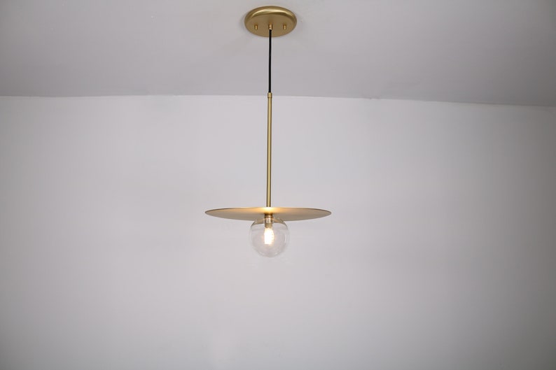 May include: A gold pendant light with a round glass shade and a flat, circular canopy. The light is hanging from a black cord.