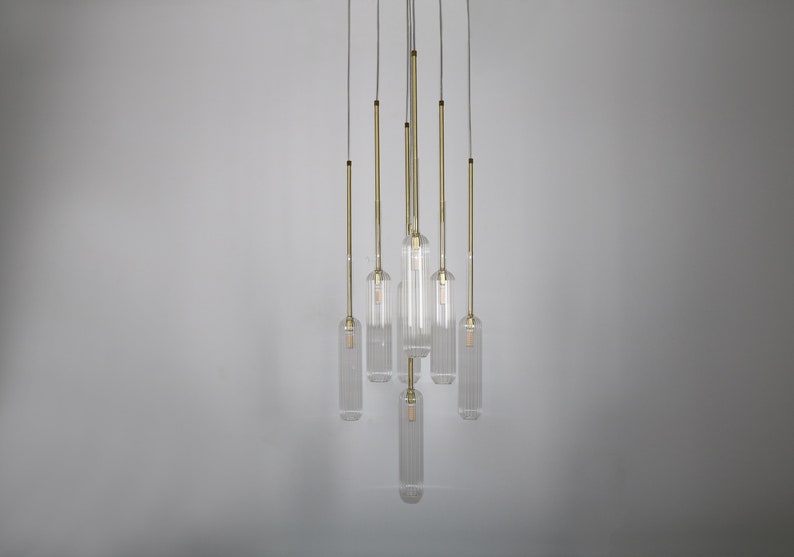 May include: A modern chandelier with seven clear glass cylinders hanging from a white ceiling. Each cylinder has a gold metal rod and a light bulb inside.