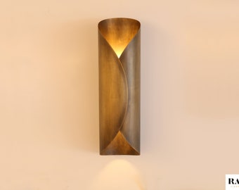 May include: A wall-mounted, brass-colored light fixture with a unique, curved design. The fixture emits a warm glow, creating a soft, inviting ambiance. The fixture is approximately 12 inches tall and is mounted on a neutral-colored wall. The brand name "RANOR" is visible at the bottom.