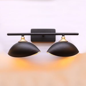 May include: Black vanity light fixture with two round shades and gold accents. The light fixture is mounted on a white wall.