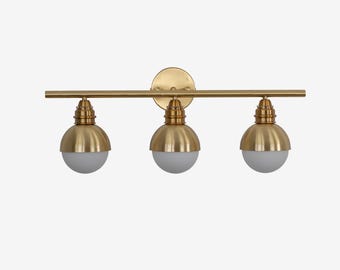 May include: A three-light vanity fixture with a gold finish. The fixture has a horizontal bar with three downward-facing lights. Each light has a gold-colored dome and a white, spherical bulb. The fixture is mounted on a round, gold backplate.