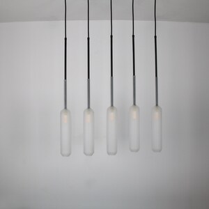 May include: Five frosted glass pendant lights hang from black cords against a white wall. The lights are cylindrical and have a ribbed texture.