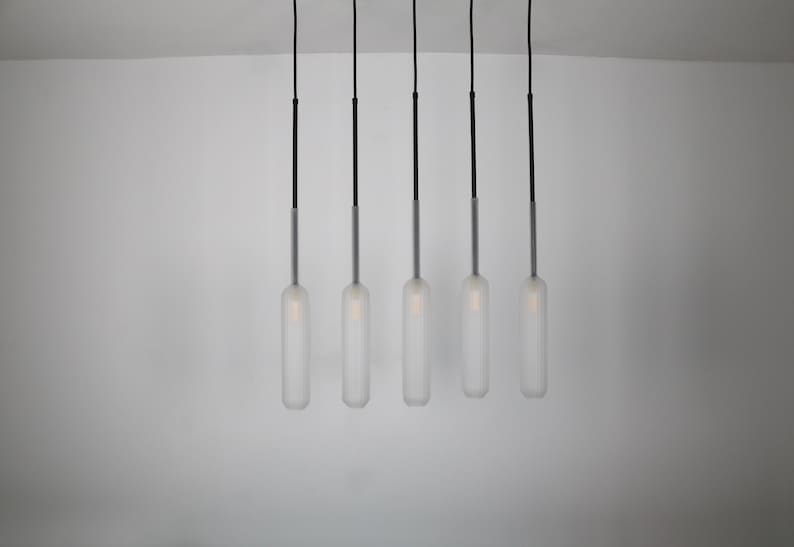 May include: Five frosted glass pendant lights hang from black cords against a white wall. The lights are cylindrical and have a ribbed texture.