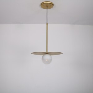 May include: A gold pendant light with a round, frosted glass shade and a flat, circular canopy. The light is suspended from the ceiling by a thin, black cord.