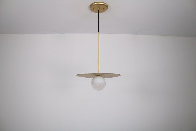 May include: A gold pendant light with a round, frosted glass shade and a flat, circular canopy. The light is suspended from the ceiling by a thin, black cord.