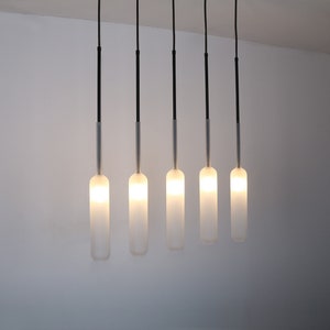 May include: Five frosted glass pendant lights hang from black cords against a white wall. The lights are turned on and emit a warm glow.