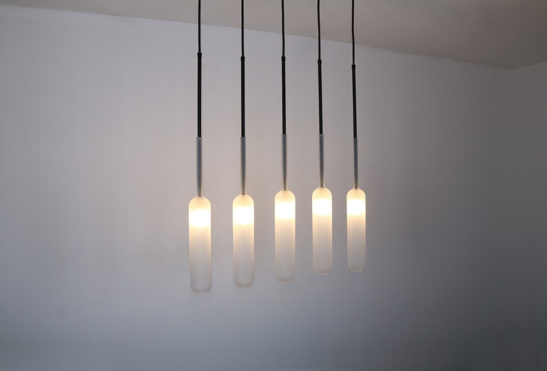 May include: Five frosted glass pendant lights hang from black cords against a white wall. The lights are turned on and emit a warm glow.
