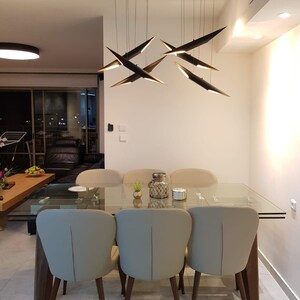 May include: A modern dining room with a glass table and six beige chairs with wooden legs. A black metal chandelier with multiple geometric light fixtures hangs above the table.