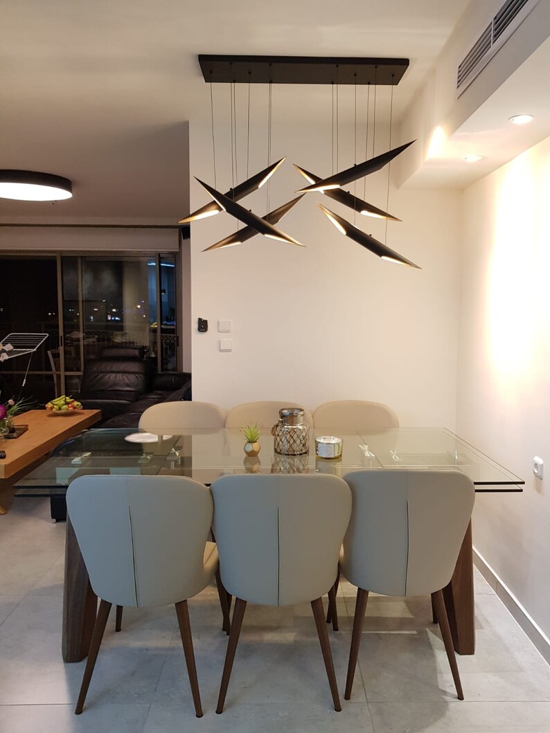 May include: A modern dining room with a glass table and six beige chairs with wooden legs. A black metal chandelier with multiple geometric light fixtures hangs above the table.