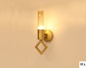 May include: Gold wall sconce with a clear glass tube and a square design. The light is on and illuminating the room.