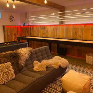 May include: A modern living room with a brown leather couch, a black pool table, and a wooden shuffleboard table. The room has a wood-paneled wall with red accent lighting and a white ceiling with a light fixture.