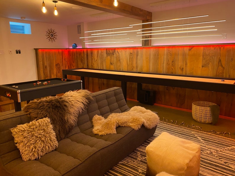 May include: A modern living room with a brown leather couch, a black pool table, and a wooden shuffleboard table. The room has a wood-paneled wall with red accent lighting and a white ceiling with a light fixture.