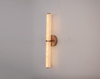 Puede incluir: Aplique de pared moderno con un diseño vertical. Cuenta con dos luminarias cilíndricas paralelas con un brillo cálido y difuso. El aplique está montado sobre una base circular de color bronce, añadiendo un toque de elegancia a la pared.